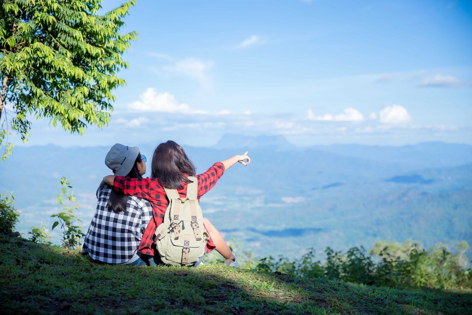 Two women sit on a hillside, one pointing at a scenic mountain landscape.
